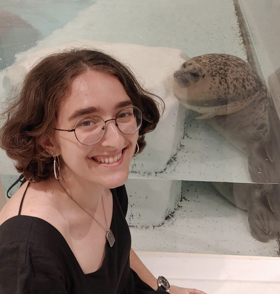 Ilana, in a black outfit, smiling and crouched by a fish tank; in the background is a seal looking very squished.